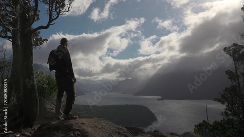 Male hiker with a backpack standing next to a tree on a high rocky viewpoint, looking out at lake during stormy, windy weather