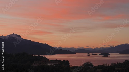 Panoramic wide shot of Lake Nahuel Huapi and hotel nestled among forested hills under a dramatic orange and pink sunset sky in Bariloche, Argentina.