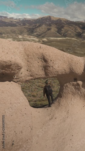 Male hiker with a backpack standing and looking through a natural stone arch hole, with a dry valley and mountain range under a cloudy sky in the background.