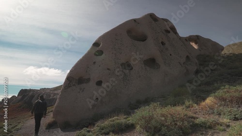 Medium shot from behind of a male tourist with a backpack standing next to a large porous stone boulder showing natural erosion on a hillside in La Buitrera, Bariloche, Argentina.