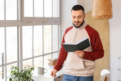 Handsome young man reading book near window at home