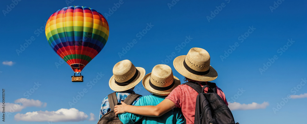 Obraz premium Three kids wear hats watch rainbow hot air balloon fly in clear blue sky. Friends with backpacks share moment of wonder on vacation. Sunny day, pure joy.