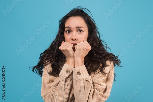 Fotografie Worried Young Woman With Dark Hair Looking Anxious And Stressed, Holding Hands To Face In Front Of Bright Blue Background