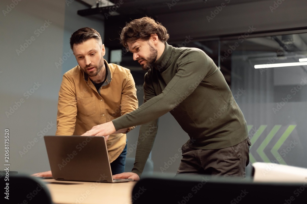 Fototapeta premium Focused Men, One Caucasian And One Middle Eastern, Collaborating On Laptop In Modern Office Setting, Discussing Project Details And Working Together Towards A Common Goal, Business Concept.