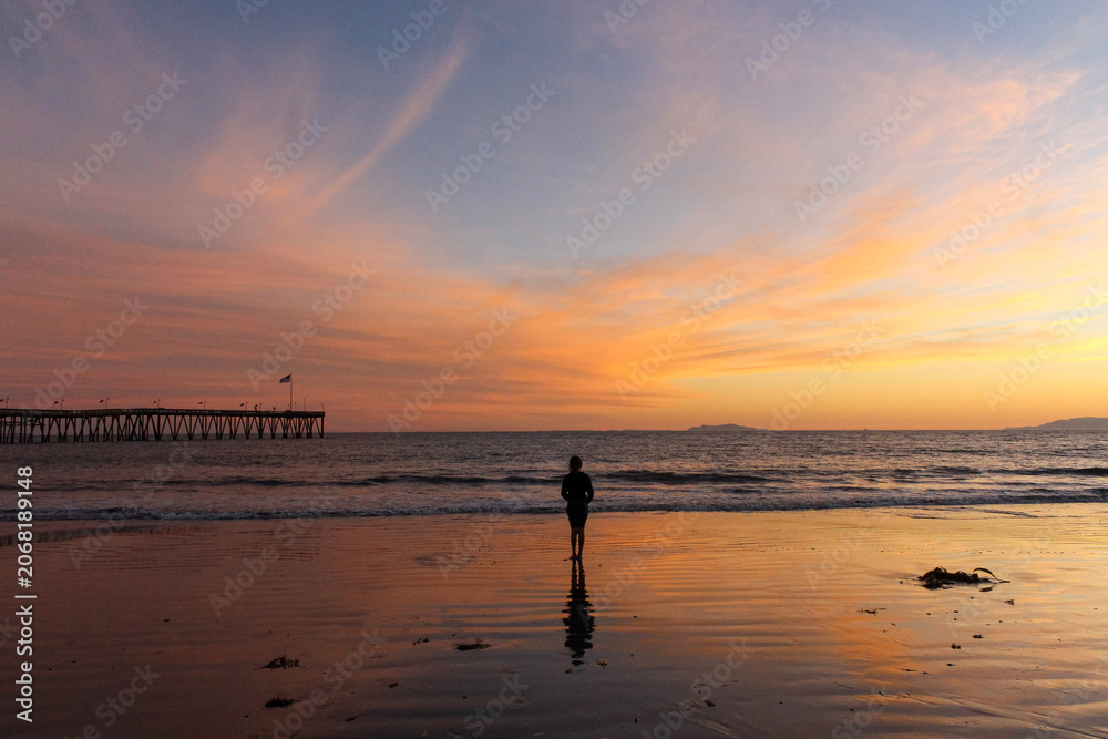 Fototapeta premium Person Standing on Beach at Colorful Sunset Near Ocean Pier