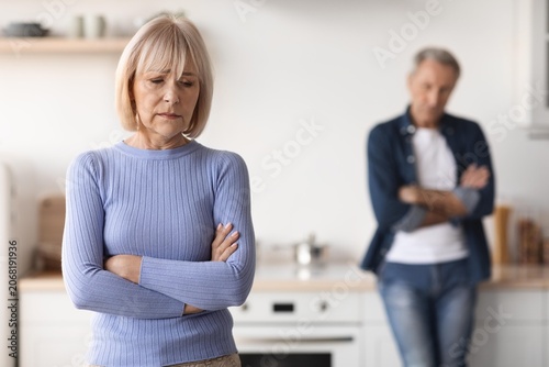 Schilderij op canvas Worried Elderly Caucasian Woman With Arms Crossed Looking Down While Elderly Man With Arms Crossed Stands In Background, Facing Relationship Conflict At Home In Kitchen