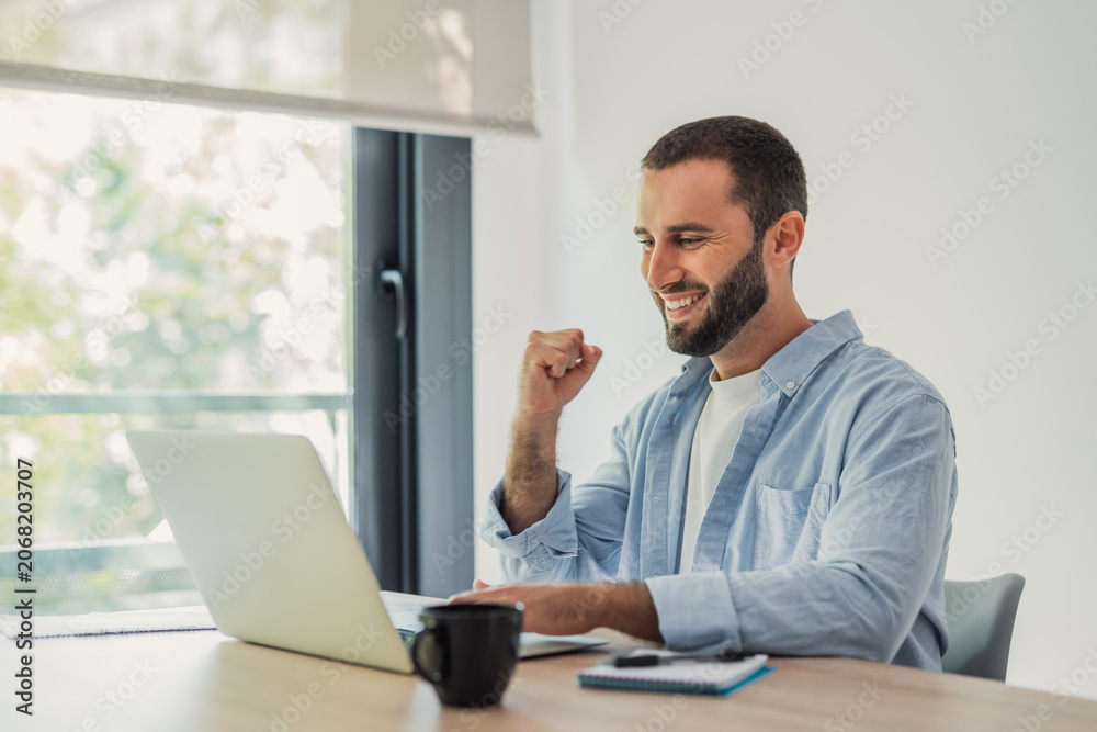 Naklejka premium Young man smiling and raising his fist in celebration while looking at his laptop screen, feeling accomplishment working remotely from a modern home office