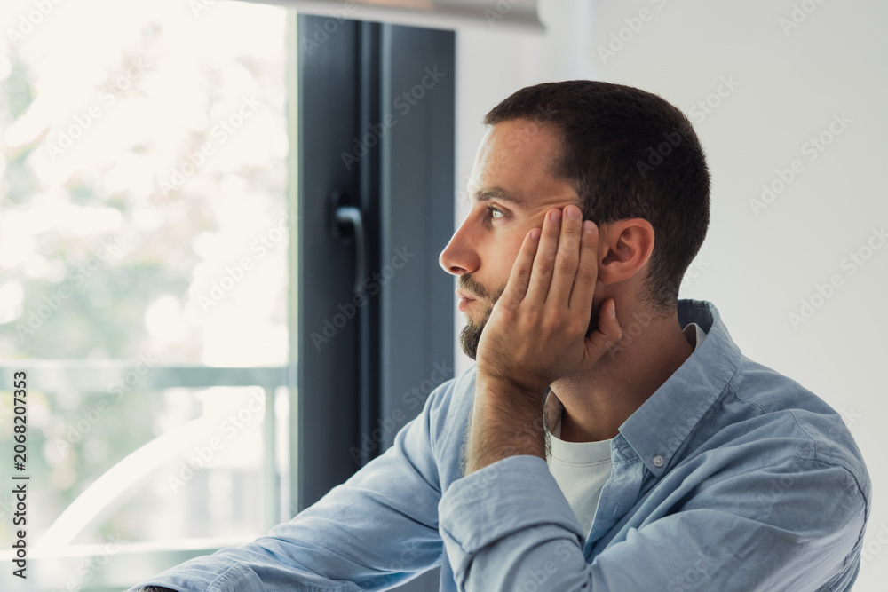 Obraz premium Young man thinking and looking away through a window. Resting his face on his hand. Reflecting on thoughts while being alone in an indoor office or home setting. Experiencing anxiety. Solitude