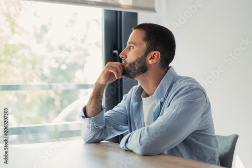 Fotografi Young man with a beard sitting at a table near a window, looking out with a thou