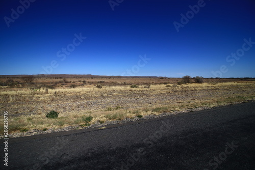 Fotografie Flat arid landscape and dry scrubland next to an asphalt road under a clear blue sky between Victoria West and Beaufort West, South Africa