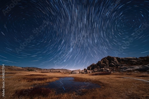 Celestial trails swirl across night sky above arid landscape with rocky outcroppings