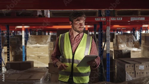 Warehouse worker inspects inventory with tablet in dimly lit warehouse setting; wearing yellow safety vest and plaid shirt while reviewing boxes and shelving units for optimal logistics.