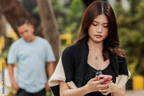 Photos A young Southeast Asian woman checks her phone, unaware that an unfamiliar man watches and trails her from behind