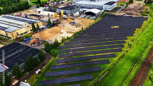 Solar panel field beside industrial buildings generating renewable energy preparatory for new build construction demand, aerial static as cloud shadows pass