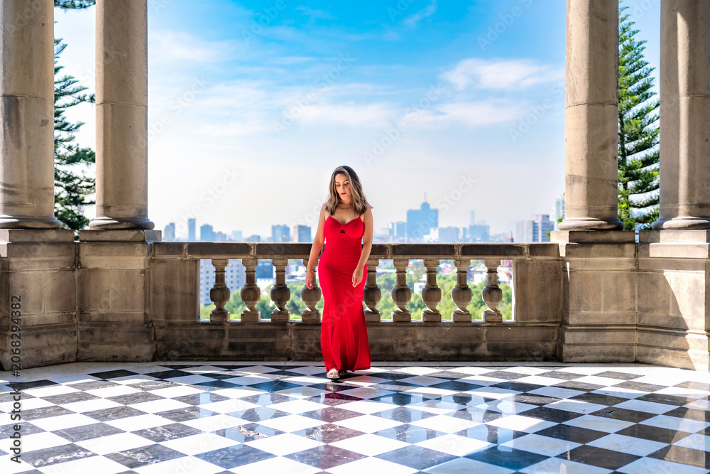 Fototapeta premium Young smiling latina woman posing confidently and calmly, enjoying the view while standing on a balcony at Castillo de Chapultepec in Mexico City, wearing an elegant long red dress
