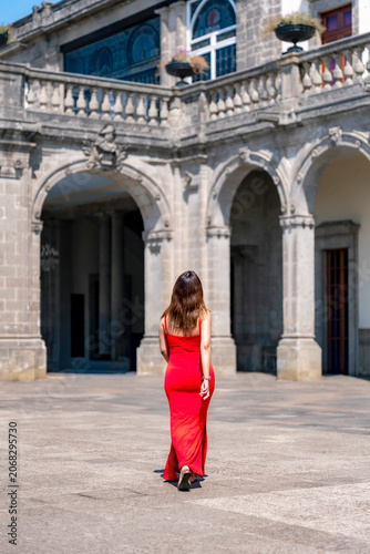 Unrecognized young latina woman walking confidently and calmly toward the museum entrance at Castillo de Chapultepec in Mexico City, wearing a vibrant fitted red dress with elegant style