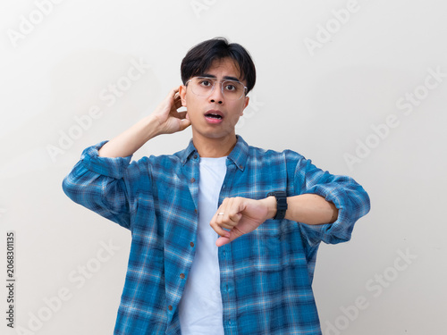 фотографія Confused young Asian man with glasses looking at his wristwatch while scratching