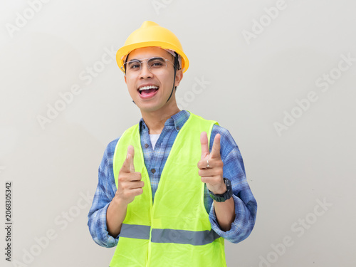 Excited young Asian engineer wearing a yellow hard hat and safety vest pointing forward to invite viewers against a plain background