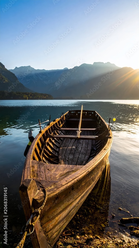Fototapeta premium Old wooden rowboat moored on a calm lake at sunrise with mountains in the background.