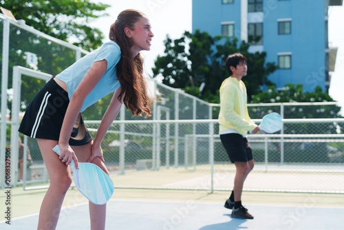 Diverse team of young caucasian woman and Asian man looking at opponent team, playing pickleball, tired and exhausted on modern outdoor court on sunny day. Sports, active and healthy lifestyle concept