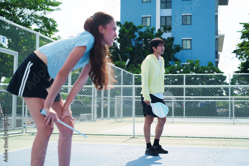 Diverse team of young caucasian woman and Asian man looking at opponent team, playing pickleball, tired and exhausted on modern outdoor court on sunny day. Sports, active and healthy lifestyle concept