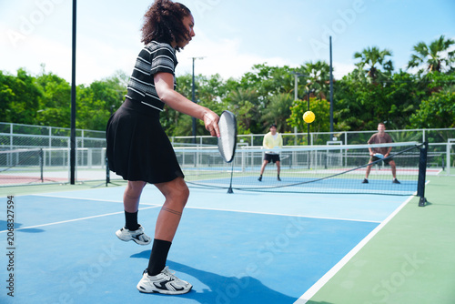 Black woman is hitting the ball, playing pickleball with a diverse group of friends in casual athletic wear on modern outdoor court on sunny day. Sports, active and healthy lifestyle concept