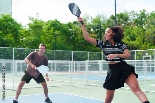 Diverse team of black woman and white man playing pickleball, woman hitting the ball back, looking at the opponent on modern outdoor court on sunny day