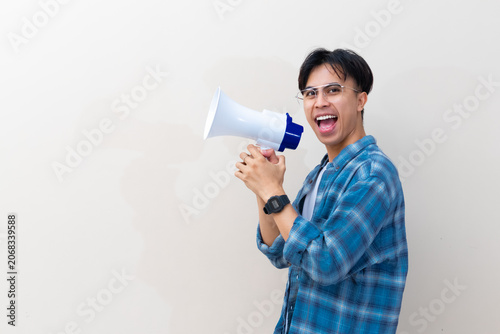 Excited young Asian man with glasses speaking into a megaphone while standing in profile view against an isolated white background