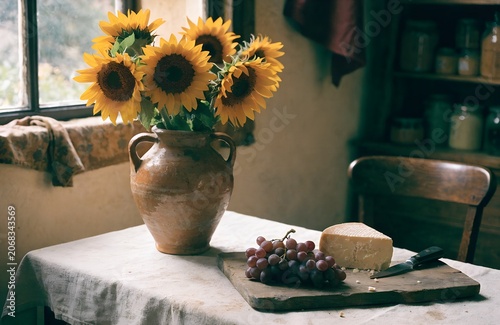 A vintage French market table with a bouquet of sunflowers in a terracotta jug beside a wooden board of dark red grapes and aged cheese soft window light faded linen