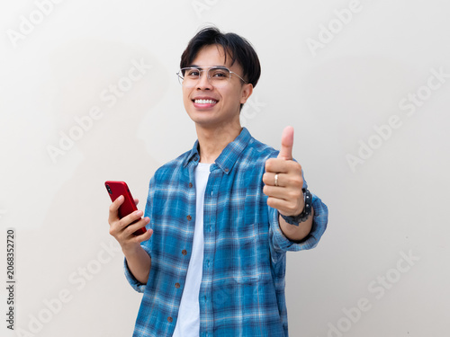 Happy young Asian man in plaid shirt holding smartphone and showing thumbs up gesture against isolated white background