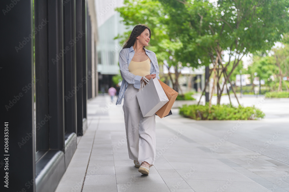 Fototapeta premium Smiling asian woman walking and holding shopping bags, Fashion shopping, retail therapy, consumerism, weekend lifestyle, purchasing power, personal reward, outdoor leisure and commercial activity