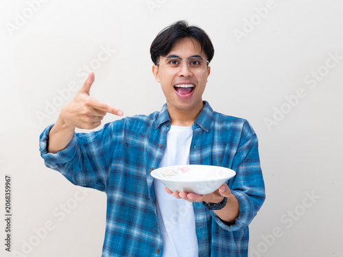 Excited young Asian man wearing glasses and a plaid shirt holds an empty plate while pointing at it against a plain background