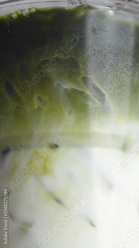 4K Vertical shot Zoom in iced matcha green tea latte in clear plastic glass on wooden table, Delicious layered beverage with rich green tea pouring into fresh white milk.