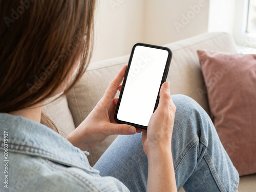 Young Woman Sitting on Sofa Holding Smartphone with Blank White Screen for Mockup App Design Presentation, Over the Shoulder View of Female Using Mobile Phone at Home
