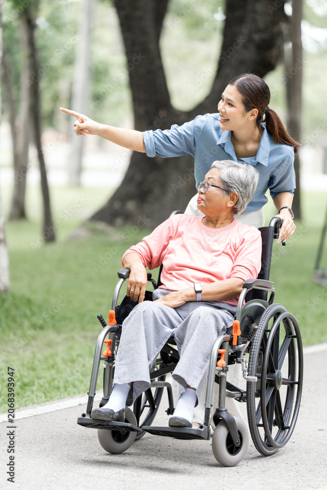 Fototapeta premium Asian Daughter Pointing Forward While Pushing Senior Mother in Wheelchair