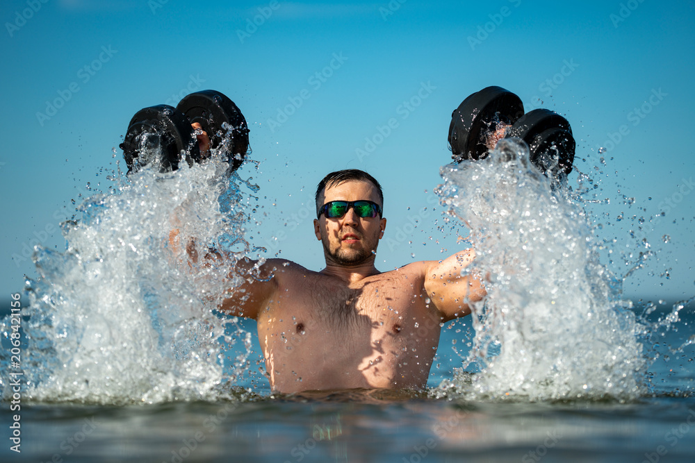Naklejka premium Dumbbells training supports summer fitness. Athlete lifts dumbbells in water. Dumbbells workout builds muscular arms. Man trains with dumbbells near waves.