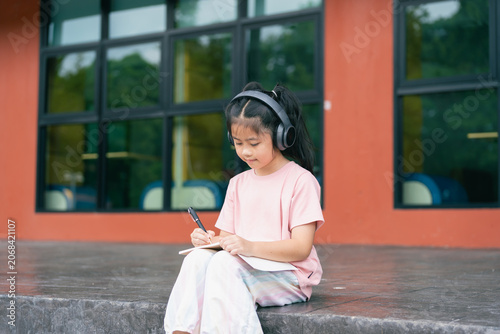 Young Girl Sitting Outdoors Wearing Headphones and Writing in Notebook Near Glass Building on a Bright Day
