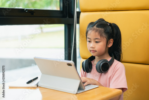 Young girl using tablet computer with headphones sitting at desk in bright classroom setting with large window and yellow seating furniture for online learning or education