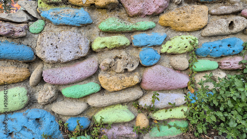 Fotografie A stone wall features porous rocks painted in vibrant shades of blue, green, pink, and yellow, with small green plants growing along the base