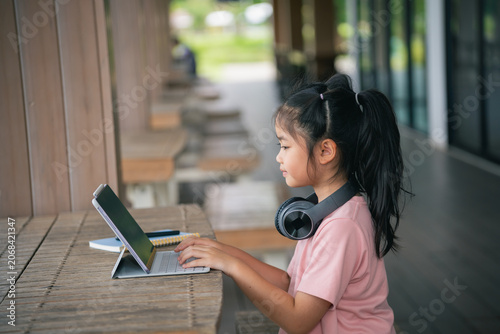Young Girl Using Laptop in Modern Indoor Space with Wooden Bench and Large Windows, Wearing Pink T-Shirt and Headphones, Enjoying Technology and Learning Environment
