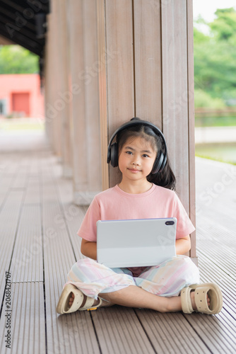 Young girl sitting on wooden floor outdoors with headphones using digital tablet, calm expression, casual pink t-shirt, colorful pants, sneakers, wooden pillars, green nature background