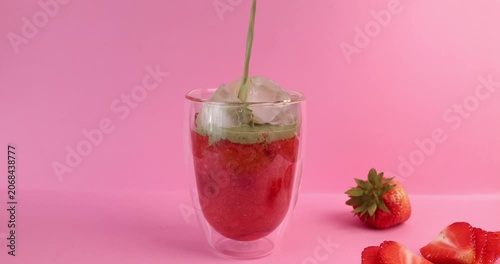 Pouring green matcha milk over strawberry puree layer in glass on pink background. Layered iced beverage concept features cold tea cocktail with ice cubes and fresh red berries. Colorful contrast shot