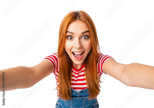 Excited redhead woman taking a selfie with a surprised expression, wearing denim overalls and a striped shirt, isolated on white.