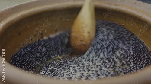 Wooden spoon stirs bubbling black mixture in clay pot, showcasing the continuous motion and texture of the cooking process in a rustic kitchen setting