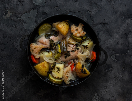 Slow cooking chicken with seasonal vegetables in a cast-iron skillet on a dark background, top view. Simple, comfort food