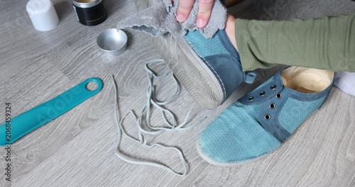Cleaning dirty sole of blue suede shoes with cloth before storage. Footwear maintenance routine concept features close up hands washing casual sneakers on bright floor background. Domestic household