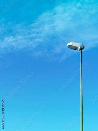 A simple yet striking urban composition. The stark form of a modern lamppost stands against a brilliant blue sky with wispy clouds, highlighting the beauty of functional city architecture.