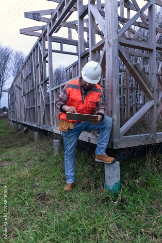 Construction worker sitting on a wooden house frame while using a digital tablet
