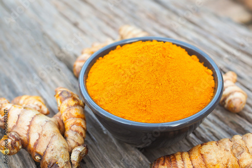 Turmeric powder in a ceramic bowl with fresh turmeric rhizomes on rustic wooden background.