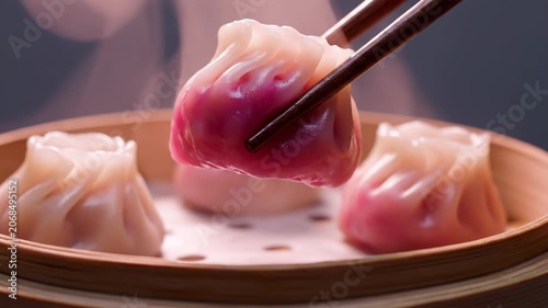Steamed dumplings with pink filling in a bamboo steamer basket. Wooden chopsticks picking up one piece with steam rising in the background. Close-up view of Asian dim sum food.